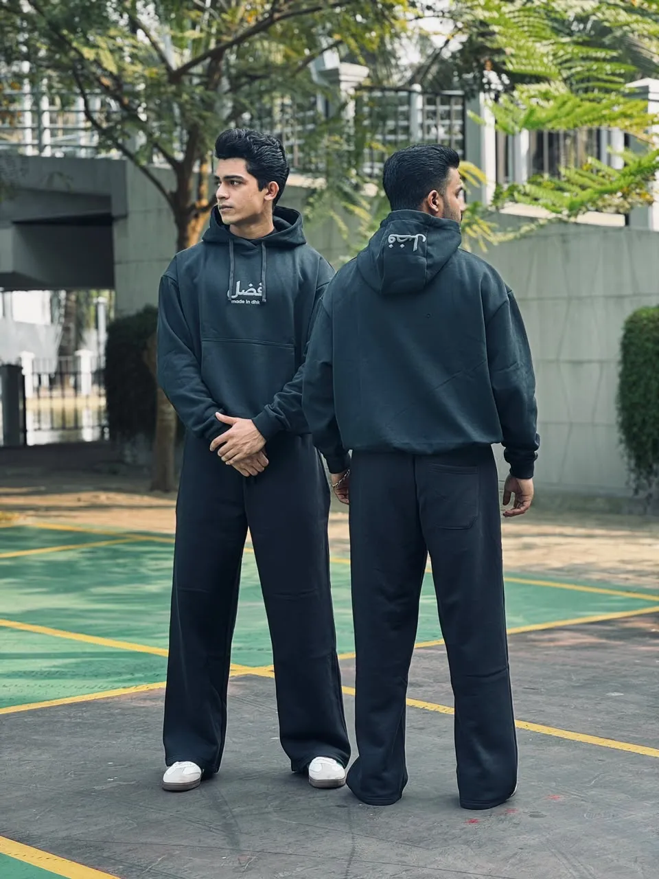 Two models wearing the black FAZAL hoodie and wide-leg trouser on an outdoor court in Dhaka