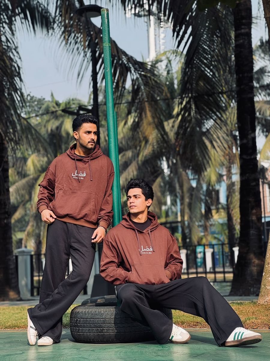 Two models in the Earth brown FAZAL hoodie and black wide-leg trouser, seated on a tyre